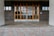 A building entrance with large glass-pane windows and two sets of double doors made of wood. The surrounding walls are made of dark, square tiles, and the floor consists of light-colored brick pavers arranged in a pattern.