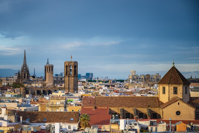 A cityscape featuring a mix of historical and modern architecture. Prominent are the spires and towers of a Gothic cathedral, possibly the Barcelona Cathedral, with its intricate designs, set against a backdrop of lower, colorful residential buildings. The skyline also includes modern structures, blending history with contemporary elements under a partly cloudy sky.
