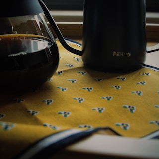 A dark-colored coffee carafe next to a sleek black kettle on a patterned yellow tablecloth. The tablecloth features small abstract designs in shades of blue and green.