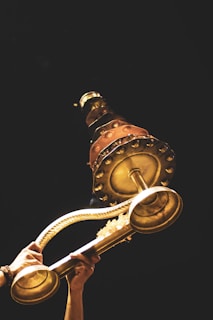 A pair of hands holds up a large, ornate brass lamp against a dark background. The lamp features intricate designs and multiple tiers, with visible floral decorations and metallic accents.