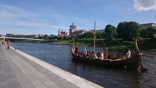 A wooden Viking-style longboat with people on board is sailing along a river bordered by a landscaped promenade. A historic cityscape with a prominent church tower and other buildings is visible on the opposite bank.
