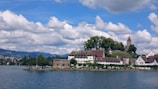 An idyllic scene features a small town situated by a lake, highlighted by historic buildings with sloping roofs and a church with a tall steeple. Lush green trees surround the area, and a cloudy sky dominates the background.