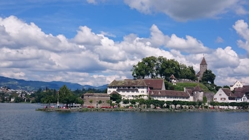 An idyllic scene features a small town situated by a lake, highlighted by historic buildings with sloping roofs and a church with a tall steeple. Lush green trees surround the area, and a cloudy sky dominates the background.