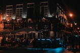A dimly lit street scene with an illuminated cafe named 'O Brasileiro'. People are seated under large parasols, engaging in conversation. The atmosphere is warm with soft lighting from street lamps.