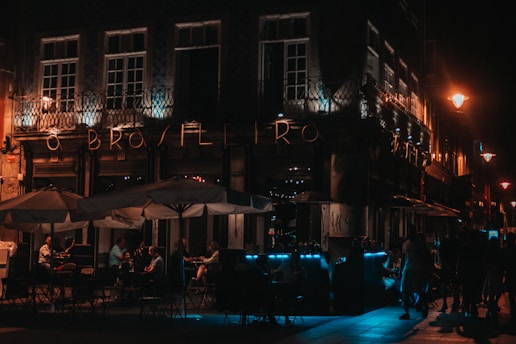 A dimly lit street scene with an illuminated cafe named 'O Brasileiro'. People are seated under large parasols, engaging in conversation. The atmosphere is warm with soft lighting from street lamps.