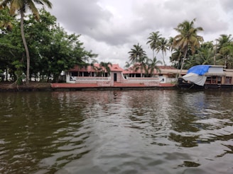 A peaceful waterfront scene with a traditional house featuring red-tiled roofs and a brick façade. Palm trees are scattered across the background, contributing to a tropical ambiance. A houseboat is moored to the side, partially covered with a blue and white tarp. The water in the foreground reflects the cloudy sky above.