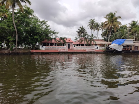 A peaceful waterfront scene with a traditional house featuring red-tiled roofs and a brick façade. Palm trees are scattered across the background, contributing to a tropical ambiance. A houseboat is moored to the side, partially covered with a blue and white tarp. The water in the foreground reflects the cloudy sky above.