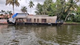 A traditional houseboat is moored along a calm body of water. The houseboat has a wooden structure with multiple windows and a partially covered top. Palm trees and lush green foliage are visible in the background alongside a building with a red tiled roof.