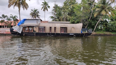 A traditional houseboat is moored along a calm body of water. The houseboat has a wooden structure with multiple windows and a partially covered top. Palm trees and lush green foliage are visible in the background alongside a building with a red tiled roof.