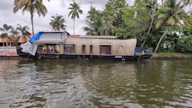 A traditional houseboat is moored along a calm body of water. The houseboat has a wooden structure with multiple windows and a partially covered top. Palm trees and lush green foliage are visible in the background alongside a building with a red tiled roof.