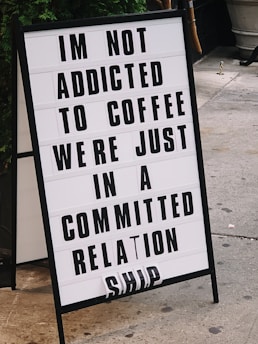 A sandwich board sign on a sidewalk displays a humorous message about coffee addiction and relationship commitment. The letters are bold and black against a white background. The board is positioned on a concrete surface with some foliage visible in the background.