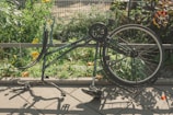 A bicycle frame is seen without its front wheel and saddle, resting upside down on a paved surface. The background features lush green plants and yellow flowers, enclosed by a metal fence. The sunlight casts distinct shadows of the bicycle onto the pavement.