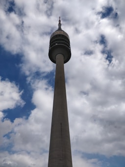A tall telecommunications tower rises into a partly cloudy sky. The tower is cylindrical with a wider section towards the top, and it has multiple rings and antennae. The sky is mostly covered with fluffy clouds against patches of blue, giving a sense of height and openness.