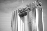 A black and white image of a high-rise building under construction with two parallel towers connected by a structure at the top. Cranes are visible, indicating ongoing construction work. The facade of the building shows a patterned design.