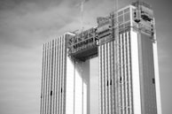 A black and white image of a high-rise building under construction with two parallel towers connected by a structure at the top. Cranes are visible, indicating ongoing construction work. The facade of the building shows a patterned design.