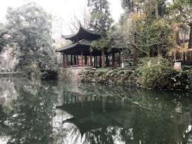 A serene scene features a traditional pagoda-style pavilion surrounded by lush greenery and towering trees. The structure is reflected in the calm, mirror-like water of a pond. The atmosphere is tranquil and harmonious, evoking a sense of peace and natural beauty.