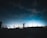 A person stands on a rooftop parking lot at night, silhouetted against a clear sky filled with stars. They hold and appear to be looking through a large camera lens or telescope. The scene is illuminated by distant city lights on the horizon, creating a gradient of blue and black in the sky.