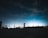 A person stands on a rooftop parking lot at night, silhouetted against a clear sky filled with stars. They hold and appear to be looking through a large camera lens or telescope. The scene is illuminated by distant city lights on the horizon, creating a gradient of blue and black in the sky.