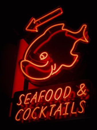 A vibrant neon sign featuring an outline of a fish and an arrow above it. Below the fish, the words 'SEAFOOD & COCKTAILS' are illuminated in glowing red neon. The sign emits a warm, inviting glow against a dark background.