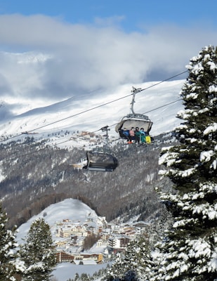 A ski resort in a mountainous region features a cable car lift with several skiers sitting and enjoying the ride. Snow-covered pine trees frame the scene, and a picturesque village is visible in the valley below. The sky is partly cloudy with patches of bright blue.