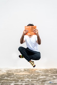 A person is levitating against a plain white background while reading an orange book. They are dressed in a white t-shirt, black pants, and colorful striped socks. The ground beneath them is made of rustic, uneven stone.