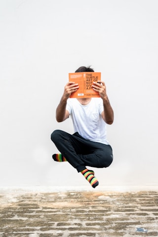 A person is levitating against a plain white background while reading an orange book. They are dressed in a white t-shirt, black pants, and colorful striped socks. The ground beneath them is made of rustic, uneven stone.