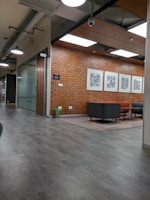A modern office space with a brick wall adorned with framed QR codes. The ceiling features large square fluorescent lights, and the floor is covered with a smooth, grey surface. In the seating area, there are black and brown couches arranged around a small table. A corridor leads to another part of the office, and there are bins positioned beside the wall.