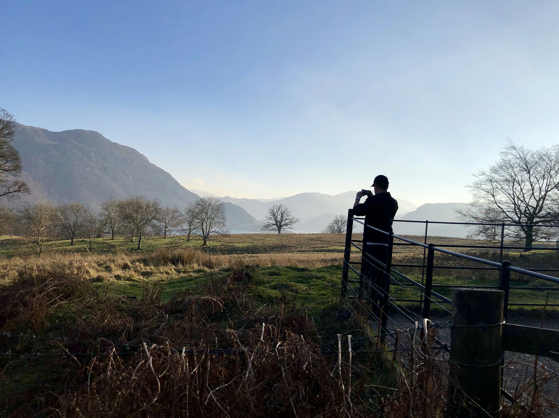A person stands on a small bridge, photographing a scenic landscape featuring rolling hills and mountains in the background. Leafless trees dot the grassy field, creating a serene and expansive view under a clear blue sky.