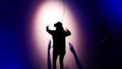 A person wearing a virtual reality headset is standing with their back to the camera, facing a wall with arms outstretched. Shadows are cast on the wall, creating a dramatic effect under a spotlight. The scene is enveloped in dark blue and purple tones, enhancing the sense of immersion and technology.