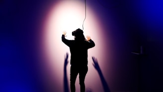 A person wearing a virtual reality headset is standing with their back to the camera, facing a wall with arms outstretched. Shadows are cast on the wall, creating a dramatic effect under a spotlight. The scene is enveloped in dark blue and purple tones, enhancing the sense of immersion and technology.