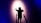 A person wearing a virtual reality headset is standing with their back to the camera, facing a wall with arms outstretched. Shadows are cast on the wall, creating a dramatic effect under a spotlight. The scene is enveloped in dark blue and purple tones, enhancing the sense of immersion and technology.