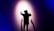 A person wearing a virtual reality headset is standing with their back to the camera, facing a wall with arms outstretched. Shadows are cast on the wall, creating a dramatic effect under a spotlight. The scene is enveloped in dark blue and purple tones, enhancing the sense of immersion and technology.