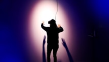 A person wearing a virtual reality headset is standing with their back to the camera, facing a wall with arms outstretched. Shadows are cast on the wall, creating a dramatic effect under a spotlight. The scene is enveloped in dark blue and purple tones, enhancing the sense of immersion and technology.
