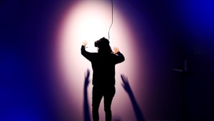 A person wearing a virtual reality headset is standing with their back to the camera, facing a wall with arms outstretched. Shadows are cast on the wall, creating a dramatic effect under a spotlight. The scene is enveloped in dark blue and purple tones, enhancing the sense of immersion and technology.