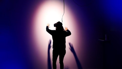 A person wearing a virtual reality headset is standing with their back to the camera, facing a wall with arms outstretched. Shadows are cast on the wall, creating a dramatic effect under a spotlight. The scene is enveloped in dark blue and purple tones, enhancing the sense of immersion and technology.
