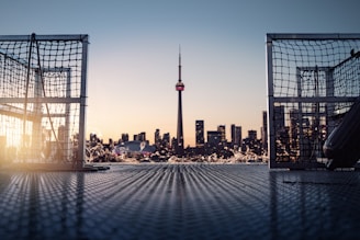 A scenic view of a city skyline with the CN Tower in the background, framed by two structures resembling hockey goals. The sunlight is casting a warm glow as it reflects off the water, creating a serene atmosphere.