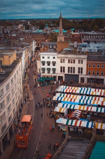 A bustling urban scene captures a busy marketplace filled with colorful vendor tents lined up in a row. People are walking around the market area, and an iconic red double-decker bus travels down a street bordered by historic buildings. The sky is overcast, adding a moody atmosphere to the scene.