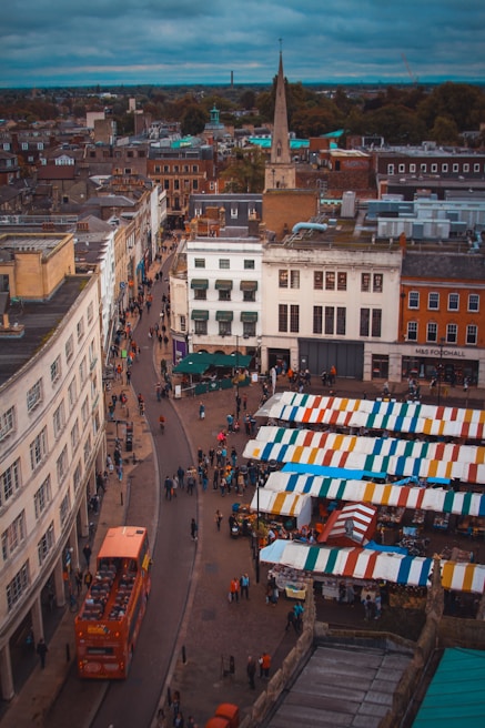 A bustling urban scene captures a busy marketplace filled with colorful vendor tents lined up in a row. People are walking around the market area, and an iconic red double-decker bus travels down a street bordered by historic buildings. The sky is overcast, adding a moody atmosphere to the scene.