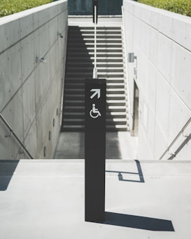 A concrete structure with a staircase leading downwards, bordered by smooth walls. A black signpost stands prominently in the foreground, featuring an arrow and a wheelchair accessibility symbol. Sunlight casts shadows on the surfaces, adding depth to the minimalist design.