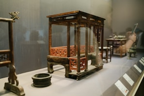 A museum exhibit featuring a wooden model of a traditional architectural structure with intricate red lattice designs. The model is raised on short legs and accompanied by a small black circular dish. In the background, other small artifacts and informational plaques are visible. A person is reflected in the glass, holding a smartphone.
