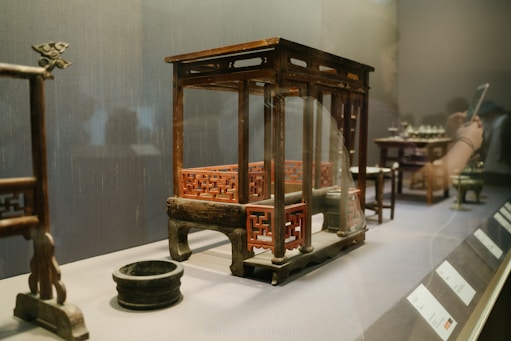 A museum exhibit featuring a wooden model of a traditional architectural structure with intricate red lattice designs. The model is raised on short legs and accompanied by a small black circular dish. In the background, other small artifacts and informational plaques are visible. A person is reflected in the glass, holding a smartphone.