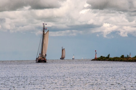 A large sailboat with prominent sails navigates an open body of water under a cloudy sky. Other smaller sailboats are visible in the distance, and a red and white striped lighthouse stands near the shore lined with greenery.