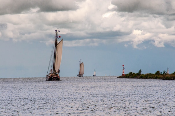 A large sailboat with prominent sails navigates an open body of water under a cloudy sky. Other smaller sailboats are visible in the distance, and a red and white striped lighthouse stands near the shore lined with greenery.