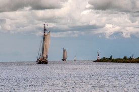 A large sailboat with prominent sails navigates an open body of water under a cloudy sky. Other smaller sailboats are visible in the distance, and a red and white striped lighthouse stands near the shore lined with greenery.