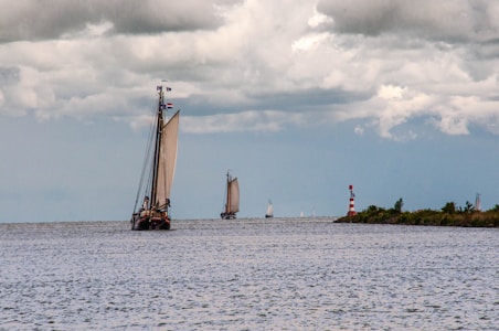 A large sailboat with prominent sails navigates an open body of water under a cloudy sky. Other smaller sailboats are visible in the distance, and a red and white striped lighthouse stands near the shore lined with greenery.