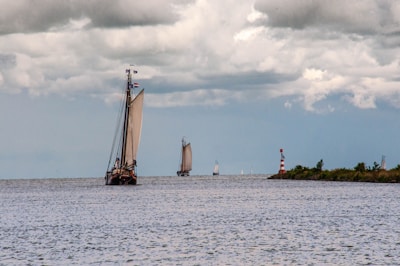 A large sailboat with prominent sails navigates an open body of water under a cloudy sky. Other smaller sailboats are visible in the distance, and a red and white striped lighthouse stands near the shore lined with greenery.