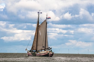 A sailing ship with tall masts and brown sails is navigating across a body of water. The sky is filled with large, fluffy clouds, and several wind turbines are visible on the horizon.