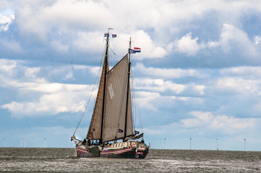 A sailing ship with tall masts and brown sails is navigating across a body of water. The sky is filled with large, fluffy clouds, and several wind turbines are visible on the horizon.