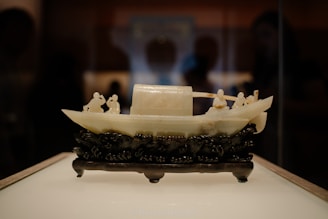 An intricately carved jade sculpture of a traditional Chinese boat, placed on a detailed black wooden base. The boat features several small figures engaged in various activities, capturing an essence of cultural artistry.