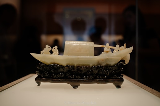 An intricately carved jade sculpture of a traditional Chinese boat, placed on a detailed black wooden base. The boat features several small figures engaged in various activities, capturing an essence of cultural artistry.